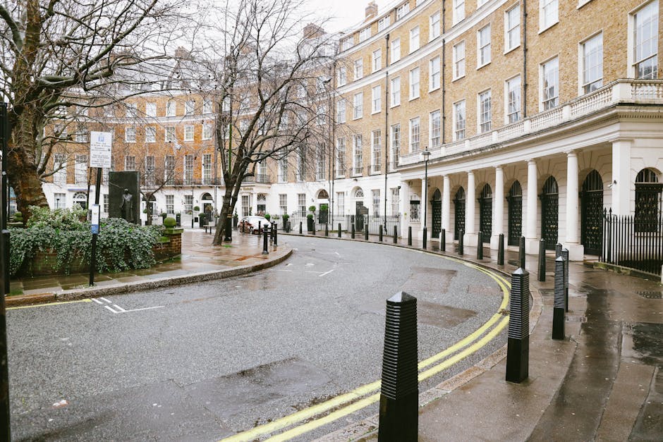 A wet street scene in Grosvenor Square, Mayfair, featuring a curved road with yellow double lines along the curb and black bollards separating the pavement from the roadway. On the left, there is a small, landscaped area with green plants, a leafless tree, and a signpost indicating parking restrictions. In the background, a row of elegant, multi-storey Georgian-style buildings with white facades, large sash windows, and black iron fences encloses the properties. The buildings have arched entrances supported by white columns, and some windows have decorative white trims. The scene appears to be during daylight hours with overcast skies, creating a subdued lighting environment. The presence of an apartment building with a flat roof and multiple windows suggests this is a residential area, where home relocation or furniture transport could involve careful loading and moving procedures by professional removal services, such as Removal Van Mayfair.