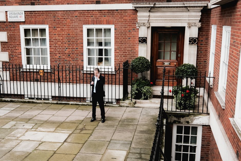 A man dressed in formal black attire, standing on a paved sidewalk outside a red-brick residential building in Mayfair. He is holding a mobile phone to his ear and appears to be communicating. The building features white-framed windows and an ornately decorated wooden door, elevated by steps with black wrought iron railings. Two potted plants with trimmed round shrubs and red flowers are positioned at the entrance. To the left, a small street sign indicates 'Grosvenor Square.' The scene suggests a home relocation or moving process, with the man possibly coordinating furniture transport or packing and moving services for the property, which is associated with Removal Van Mayfair.