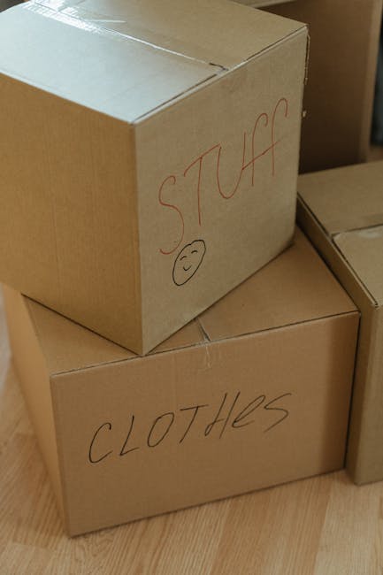 Close-up view of two plain brown cardboard boxes placed on a wooden floor inside a property, with the larger box on top labeled 'STUFF' in red marker and featuring a small smiley face, and the smaller box below labeled 'CLOTHES' in black marker. Additional boxes are partially visible nearby. The boxes are part of a home relocation packing process, reflecting careful organisation for a house move. The setting appears to be an interior space, possibly a living room or hallway, with warm lighting. These boxes are ready for transportation by Removal Van Mayfair, illustrating typical packing and loading procedures involved in furniture transport and house removals.