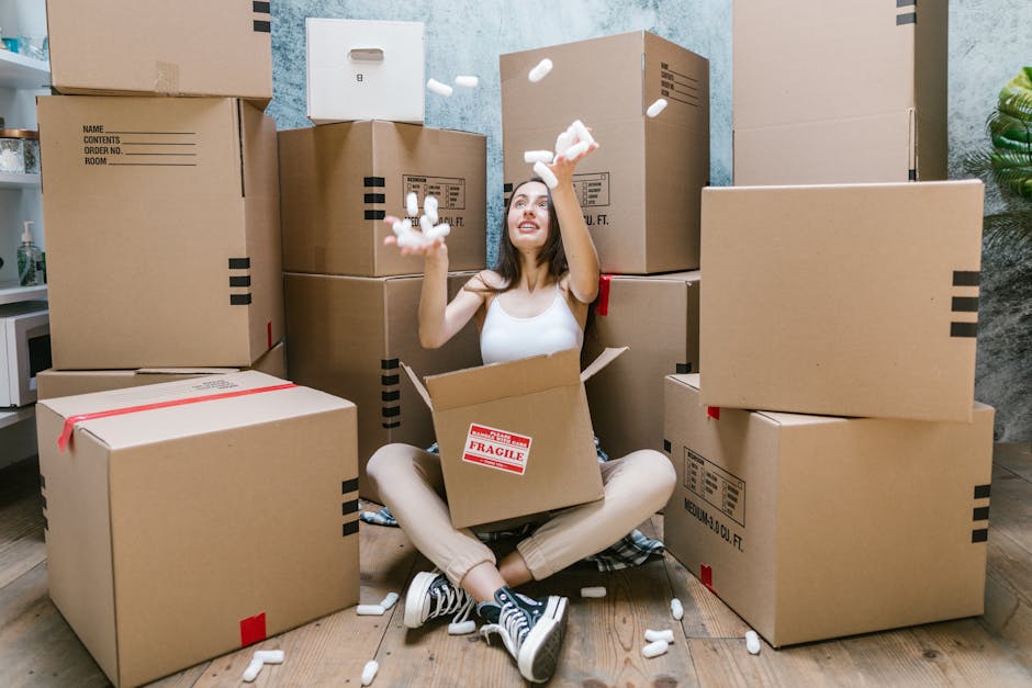 A woman sitting on a wooden floor inside a residence, surrounded by numerous large cardboard moving boxes of various sizes, some sealed with red or black tape and labeled. She is wearing a white tank top, light-colored pants, and sneakers, with a box in her lap and an open box in front of her. She is happily throwing white packing peanuts into the air, with some falling around her and others inside a partially filled box. The background shows a textured wall in a light blue or grey hue, and there is a small window or light source illuminating the scene evenly. This setting depicts a home relocation process involving packing, with furniture and personal belongings prepared for transport, supported by the services of Removal Van Mayfair for packing and moving in the Mayfair area.