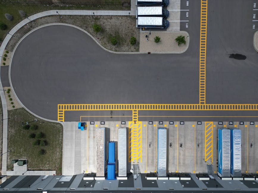 An aerial view of a busy urban street showing a parking area filled with numerous parked cars, including sedans, hatchbacks, and small SUVs, adjacent to a modern office building with large glass windo